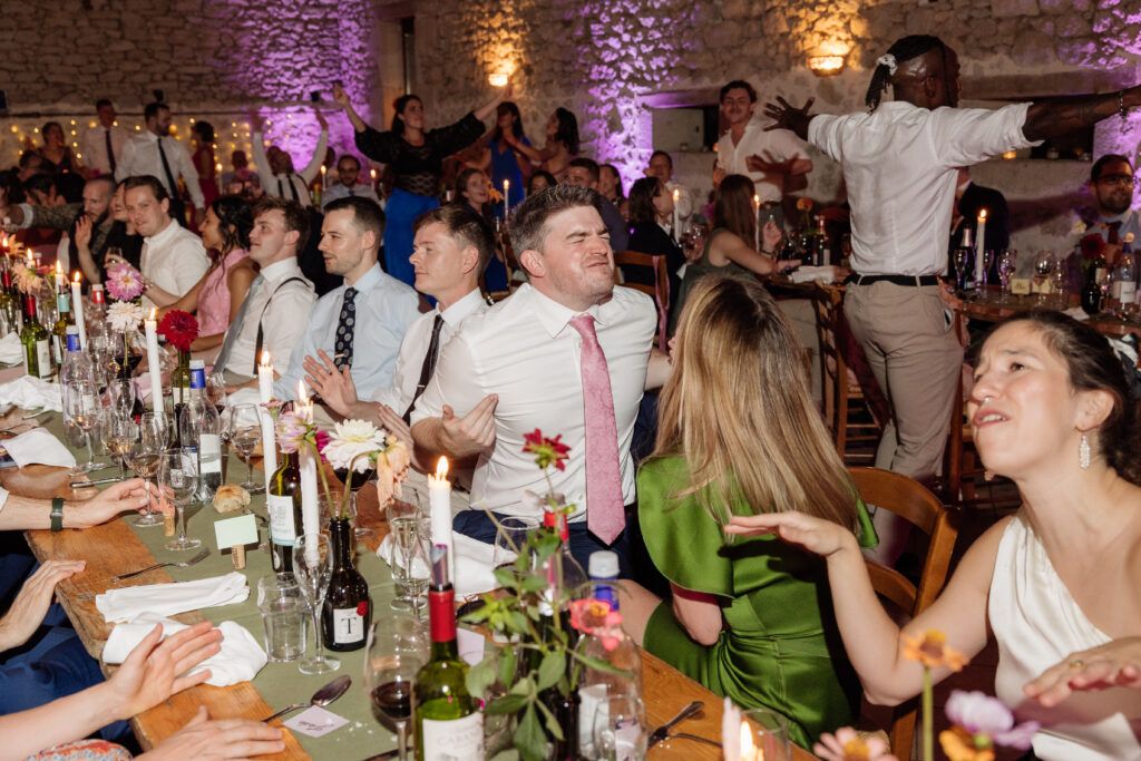 Les invités attablés lèvent les bras pendant le repas de mariage, DJ Lexvolution au fond de la grange
