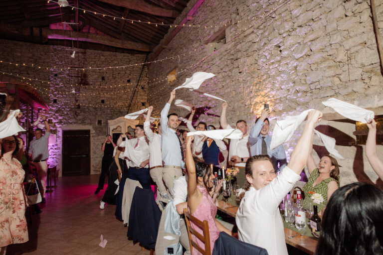 Les invités agitent leurs serviettes au-dessus des tables pendant le repas de mariage, dans une grange en pierre