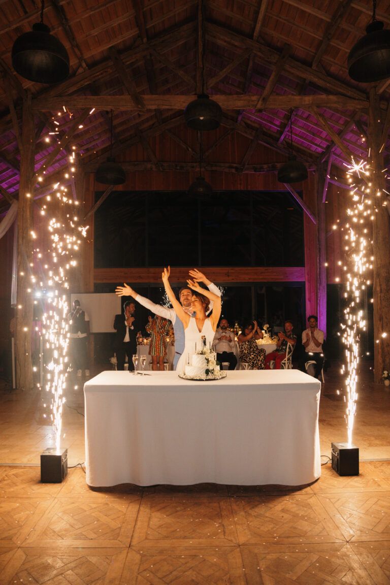 Pièce montée encadrée par deux fontaines à étincelles froides sous la charpente d'une grange, la mariée bras levés