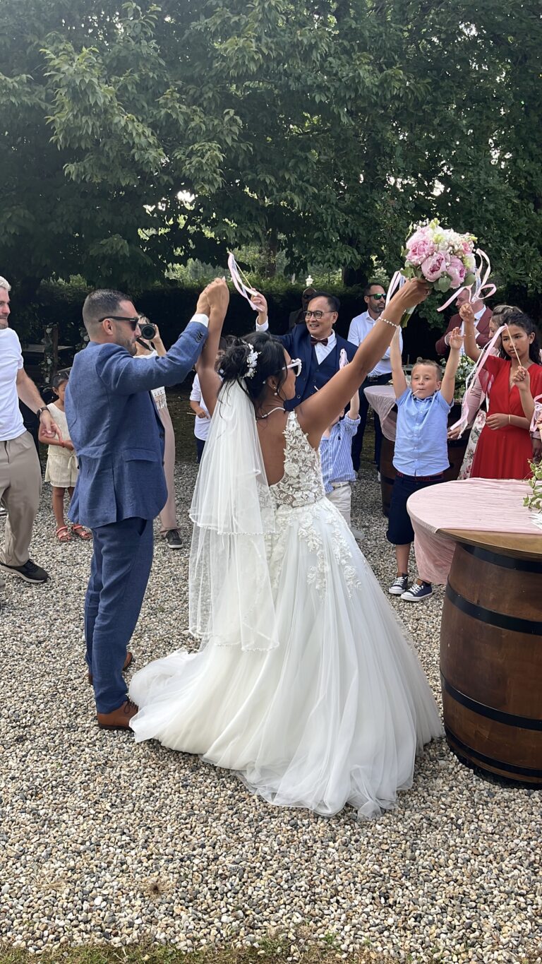 Les mariés bras levés avec le bouquet pendant le vin d’honneur en extérieur, invités autour d’un tonneau